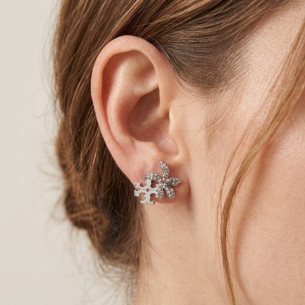 A close-up of a person's ear wearing a silver-toned stud earring with three small snowflake-like clusters set with clear stones, against a soft light beige background.