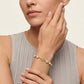 A close-up of a model wearing a delicate gold bracelet with a central monogram logo and small white pearls, paired with a light gray pleated top against a warm off-white background.