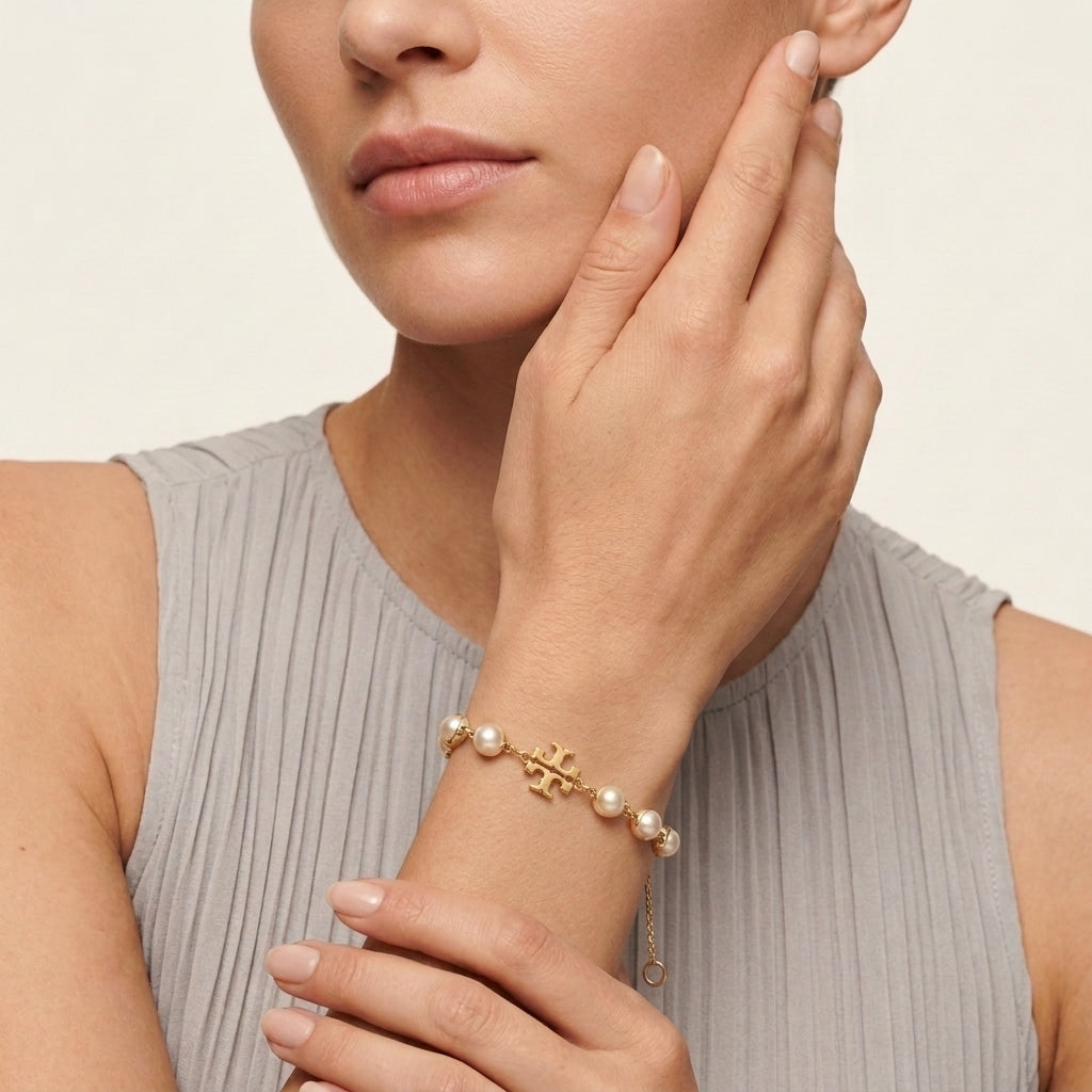 A close-up of a model wearing a delicate gold bracelet with a central monogram logo and small white pearls, paired with a light gray pleated top against a warm off-white background.