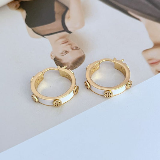 Flat lay of 3–4 different multicolour Tory Burch earring styles (e.g., enamel Kira studs, crystal-accented drops, floral motifs) on a white or light grey background.