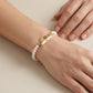 A close-up of a delicate pearl bracelet with a gold toggle clasp worn on a person's wrist, with their other hand resting nearby against a neutral beige background.