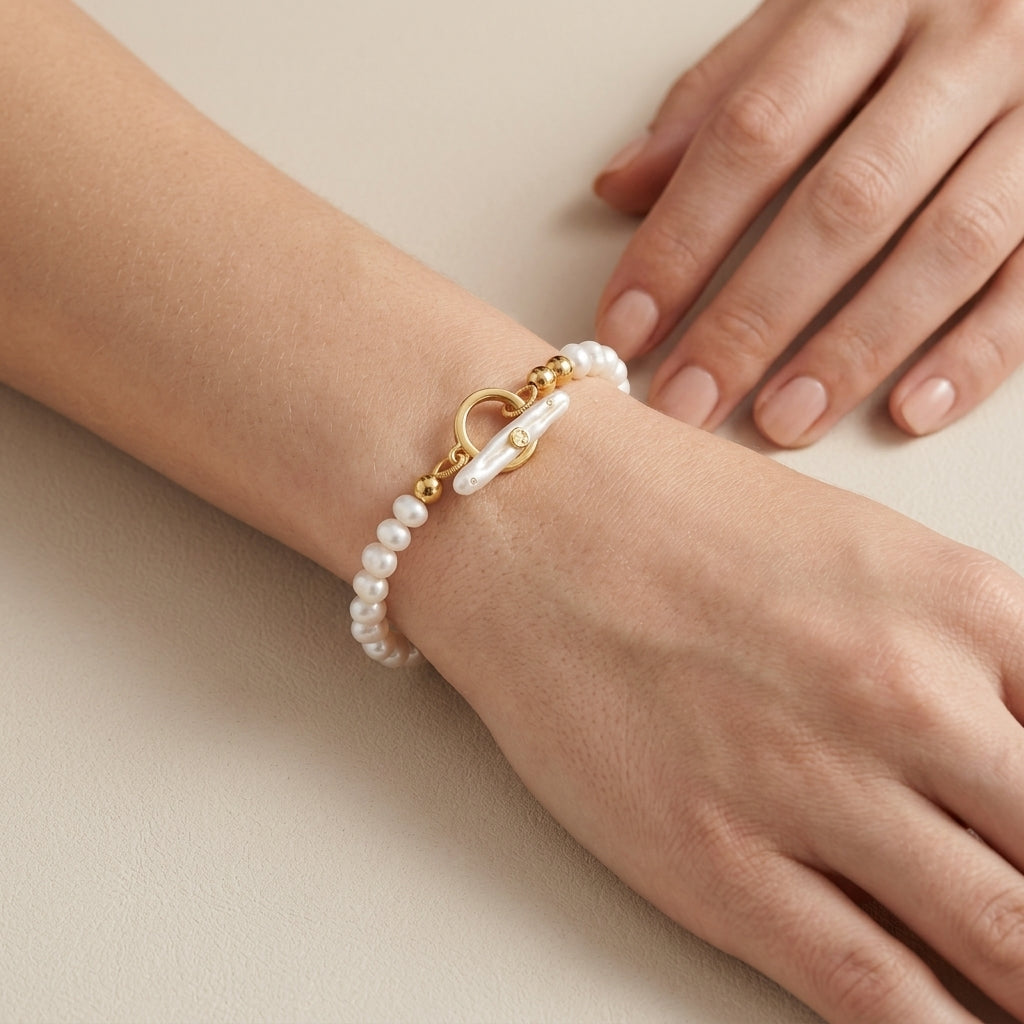 A close-up of a delicate pearl bracelet with a gold toggle clasp worn on a person's wrist, with their other hand resting nearby against a neutral beige background.