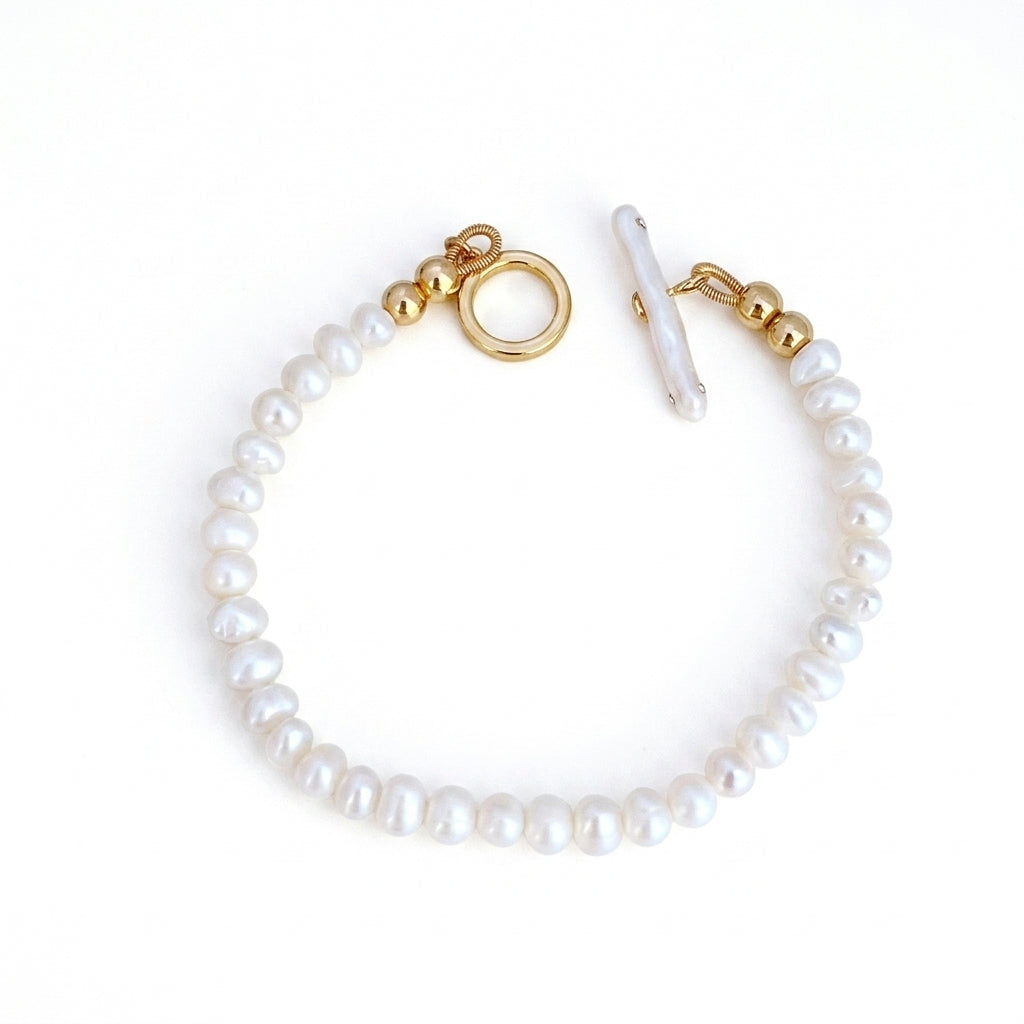 A delicate bracelet made of white baroque pearls and gold-toned metal hardware, featuring a toggle clasp and a round ring accent, shown on a plain white background.