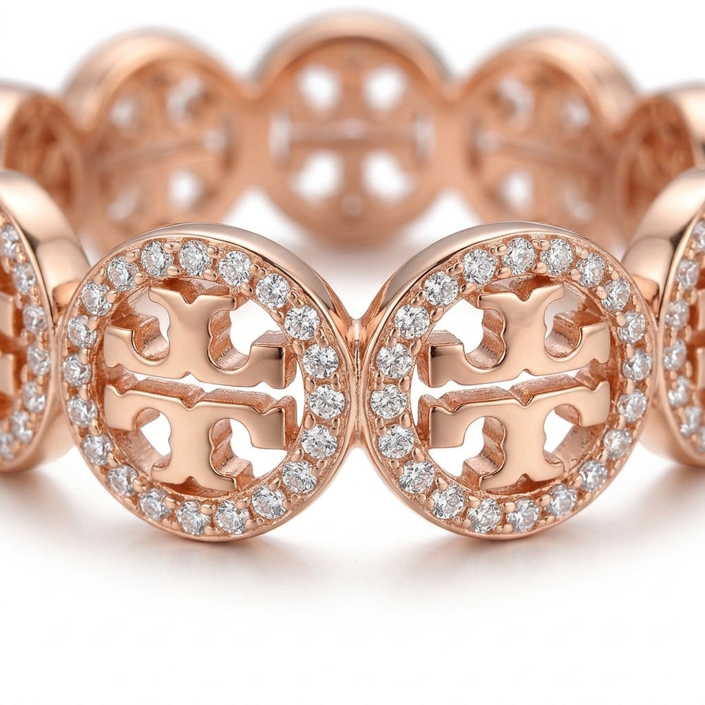 A close-up of a rose gold band featuring the interlocking Tory Burch logo surrounded by a halo of small clear stones, set against a clean white background.