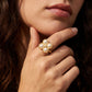 A close-up of a woman's hand wearing a gold ring with five small white pearls arranged in a flower shape, set against her chin and hair.