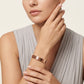 A close-up of a model wearing a slim rose gold bangle bracelet with a textured geometric pattern, paired with a light gray pleated top against a warm off-white background.