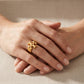 A close-up of crossed hands wearing a textured gold ring with a floral or snowflake-like openwork design, set against a soft beige fabric background.