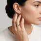 A close-up lifestyle shot of a woman wearing a small gold stud earring with a central black heart and a wide gold ring on her finger.