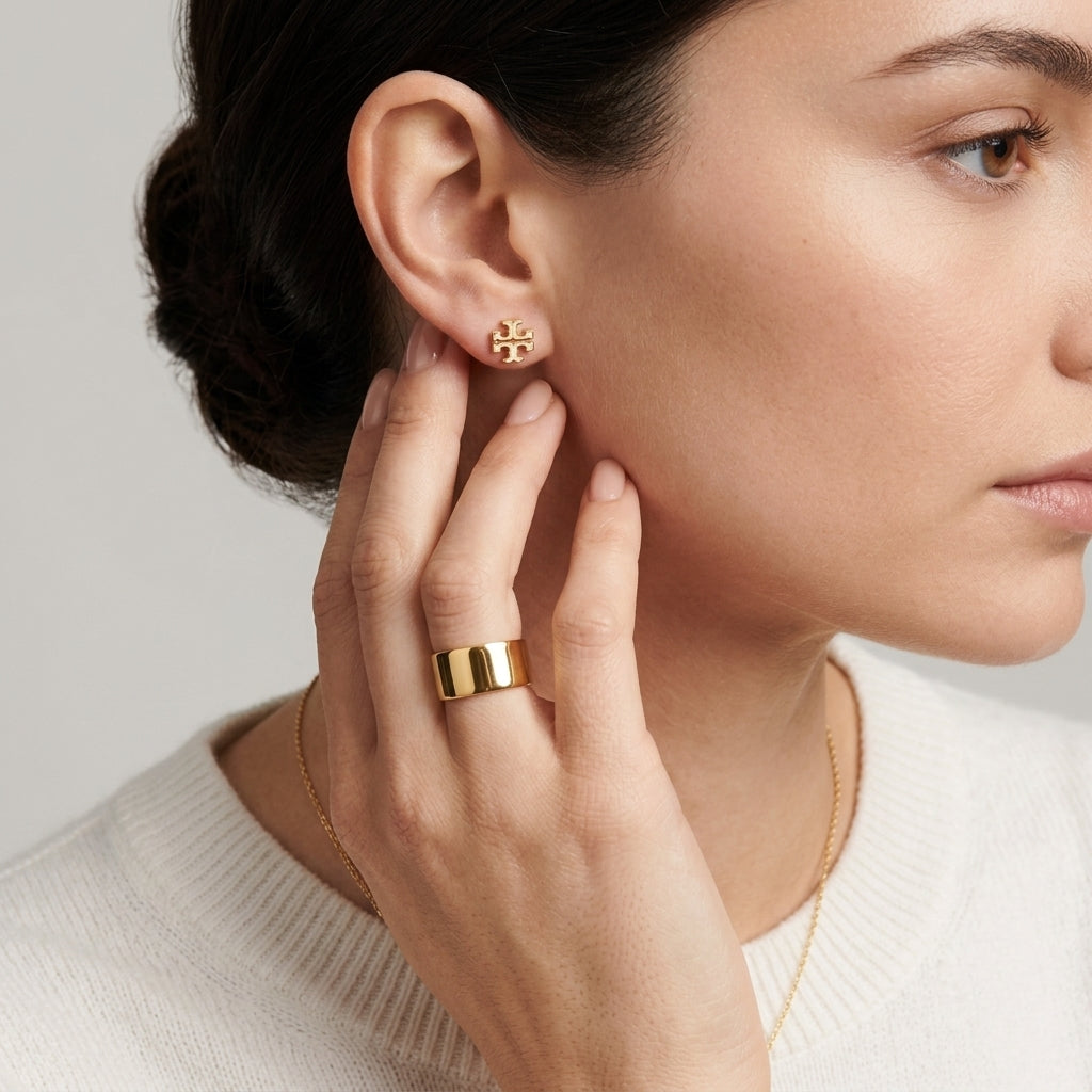 A close-up lifestyle shot of a woman wearing a small gold stud earring with a central black heart and a wide gold ring on her finger.