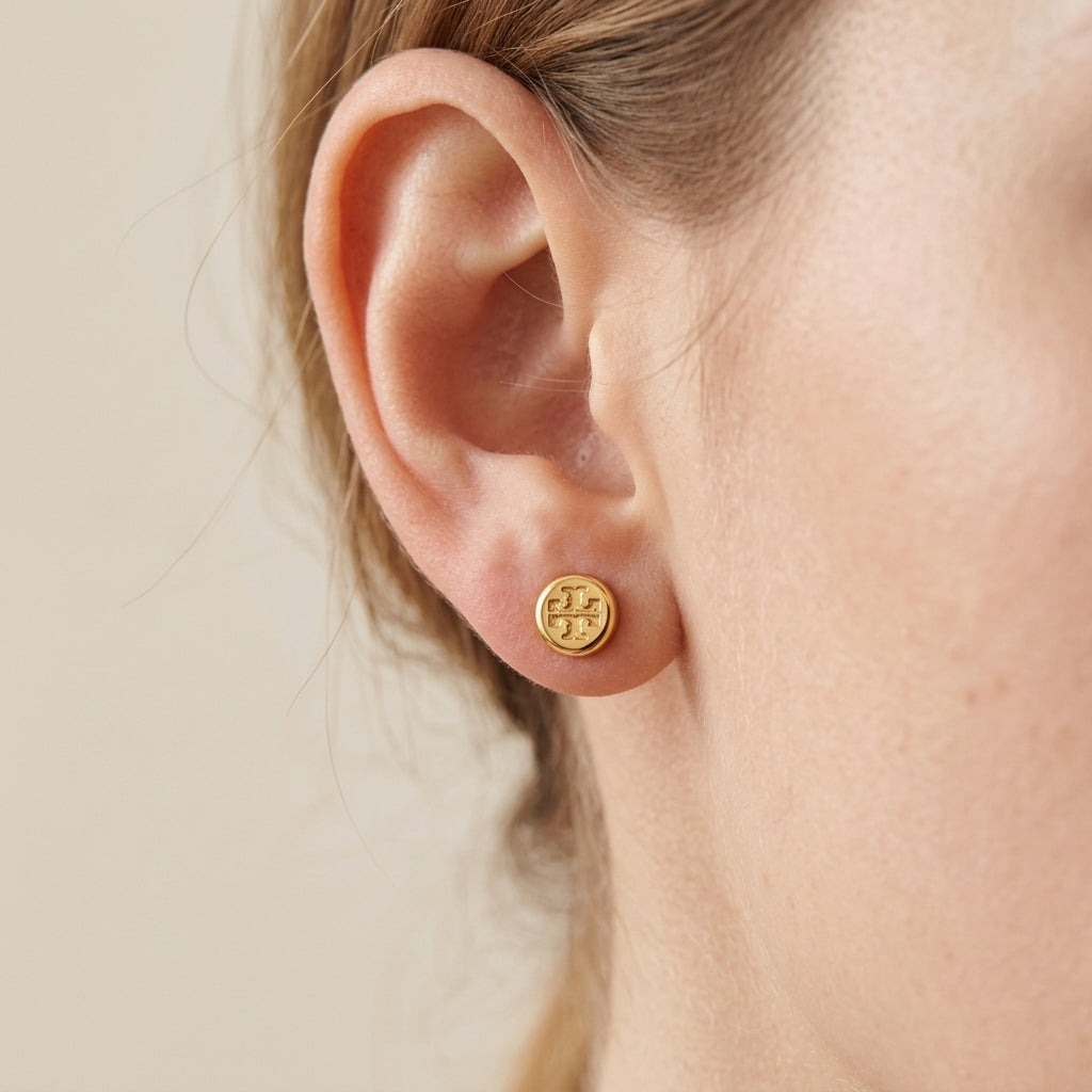 A close-up of a person's ear wearing a small round gold stud earring with an embossed monogram logo, against a soft beige background.
