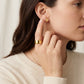A close-up lifestyle shot of a woman wearing a textured gold ring on her finger and a small gold stud earring on her ear, wearing a cream-colored ribbed sweater against a soft, light background.