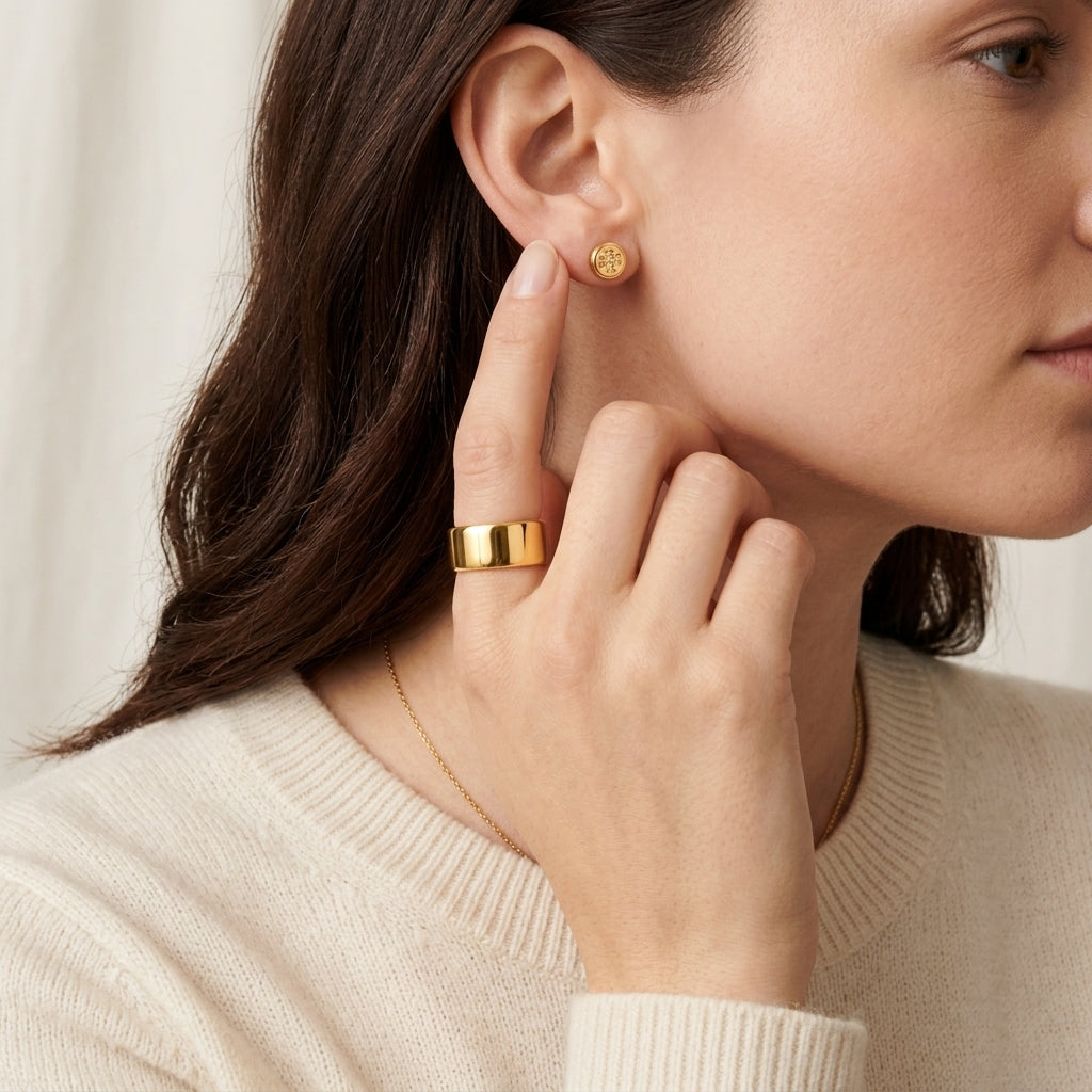 A close-up lifestyle shot of a woman wearing a textured gold ring on her finger and a small gold stud earring on her ear, wearing a cream-colored ribbed sweater against a soft, light background.