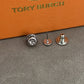 A close-up of a small metallic accessory set on a textured gray surface with an orange box branded TORY BURCH in the background. The product appears to be a pair of silver-toned stud earrings or pin-back jewelry components shown front and back.