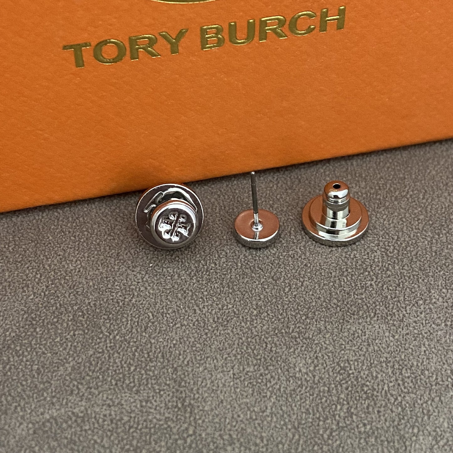 A close-up of a small metallic accessory set on a textured gray surface with an orange box branded TORY BURCH in the background. The product appears to be a pair of silver-toned stud earrings or pin-back jewelry components shown front and back.