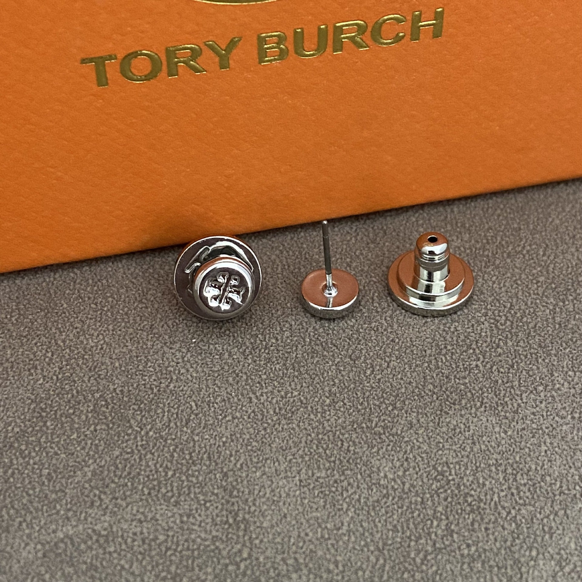 A close-up of a small metallic accessory set on a textured gray surface with an orange box branded TORY BURCH in the background. The product appears to be a pair of silver-toned stud earrings or pin-back jewelry components shown front and back.
