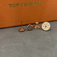 Three Tory Burch stud earrings displayed on a gray surface with an orange box in the background. The earrings include a flat round gold-tone piece with an engraved logo, a gold-tone post-back piece, and a rose-gold-tone post-back piece.