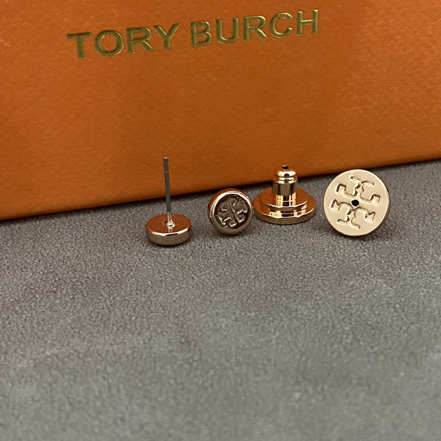 Three Tory Burch stud earrings displayed on a gray surface with an orange box in the background. The earrings include a flat round gold-tone piece with an engraved logo, a gold-tone post-back piece, and a rose-gold-tone post-back piece.
