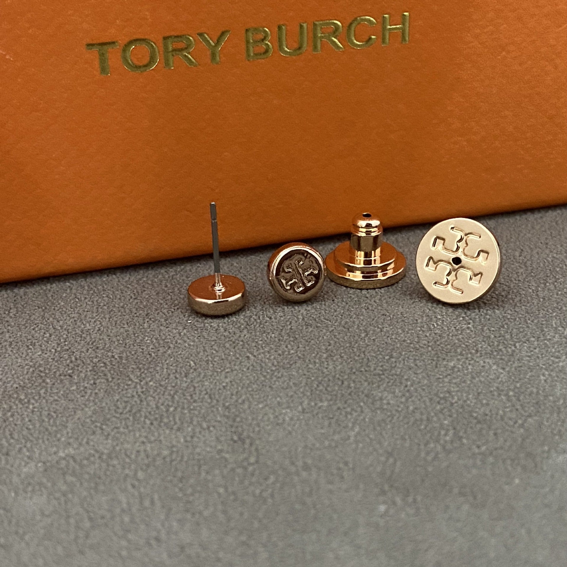 Three Tory Burch stud earrings displayed on a gray surface with an orange box in the background. The earrings include a flat round gold-tone piece with an engraved logo, a gold-tone post-back piece, and a rose-gold-tone post-back piece.