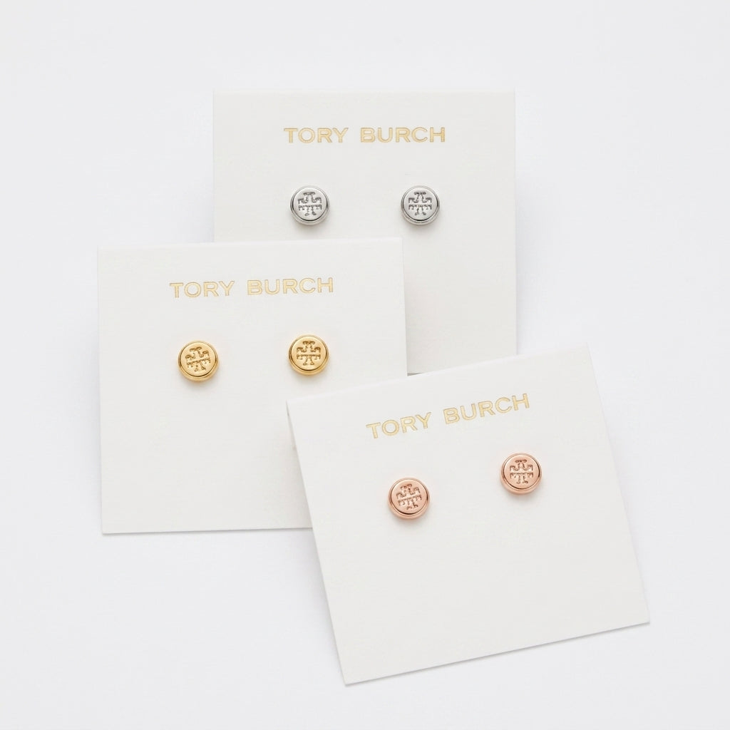 Three pairs of Tory Burch stud earrings displayed on white brand cards against a clean white background. The set includes yellow gold, silver, and rose gold options, each with a circular logo motif.