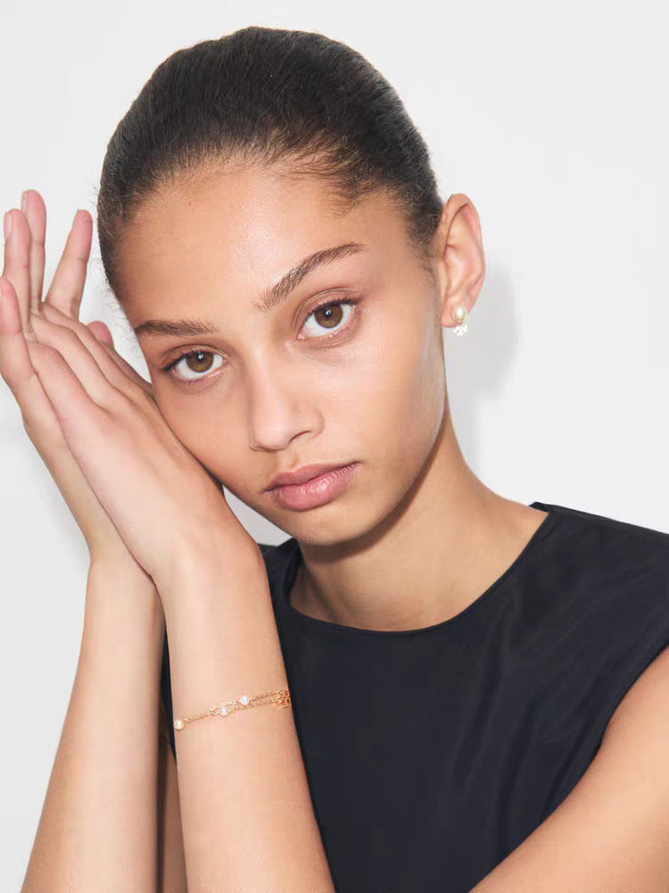 A close-up portrait of a model wearing a delicate rose gold bracelet on her wrist and a small matching stud earring, set against a clean light gray background.