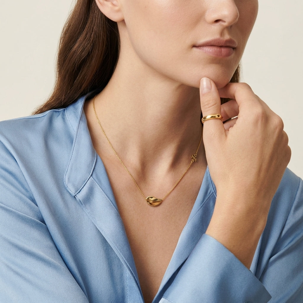 A close-up of a woman wearing a delicate gold necklace with a small curved pendant and a matching gold ring, styled with a light blue satin blouse against a soft beige background.