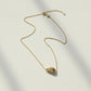 A delicate gold chain necklace with a small, polished gold leaf pendant, laid out on a light off-white surface with soft shadows.