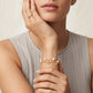 A close-up of a model wearing a delicate gold bracelet with a central gold monogram charm and small white pearls, paired with a matching gold ring on her finger.