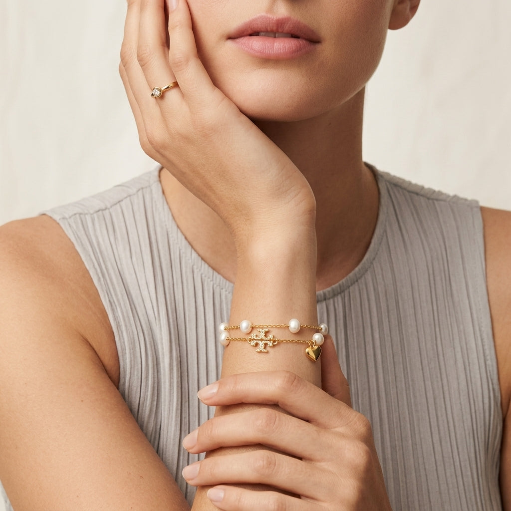 A close-up of a model wearing a delicate gold bracelet with a central gold monogram charm and small white pearls, paired with a matching gold ring on her finger.