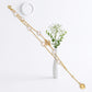 A double-strand gold chain necklace with white faux pearls, a gold flower charm, and a dangling gold heart, displayed on a white surface next to a small white vase with baby's breath.