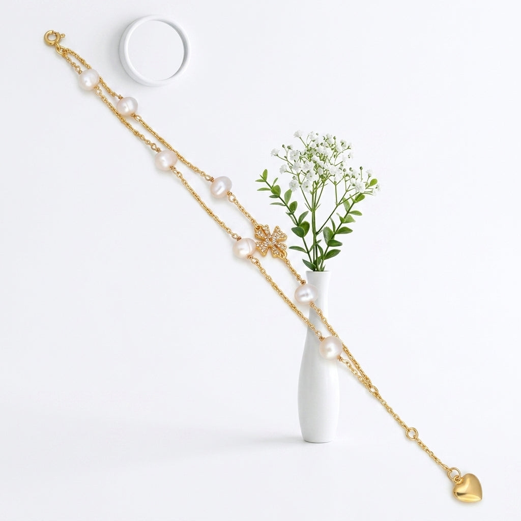 A double-strand gold chain necklace with white faux pearls, a gold flower charm, and a dangling gold heart, displayed on a white surface next to a small white vase with baby's breath.