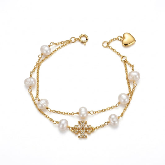A gold-tone double-strand chain bracelet featuring white freshwater pearls, a central pavé cross charm, and an adjustable heart-shaped clasp, shown on a plain white background.