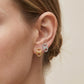 Close-up of a person's ear wearing two small stud earrings shaped like open-top bottle caps, one gold-toned and one silver-toned, against a plain light beige background.