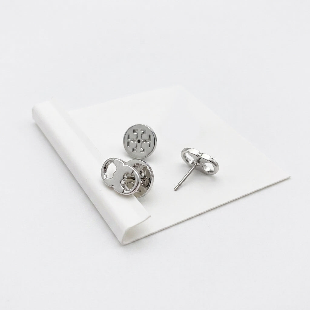 A pair of silver-toned stud earrings displayed on a white folded card against a clean white background. One earring is shown from the front with a circular cutout design, and the other is turned to show the post and backing.