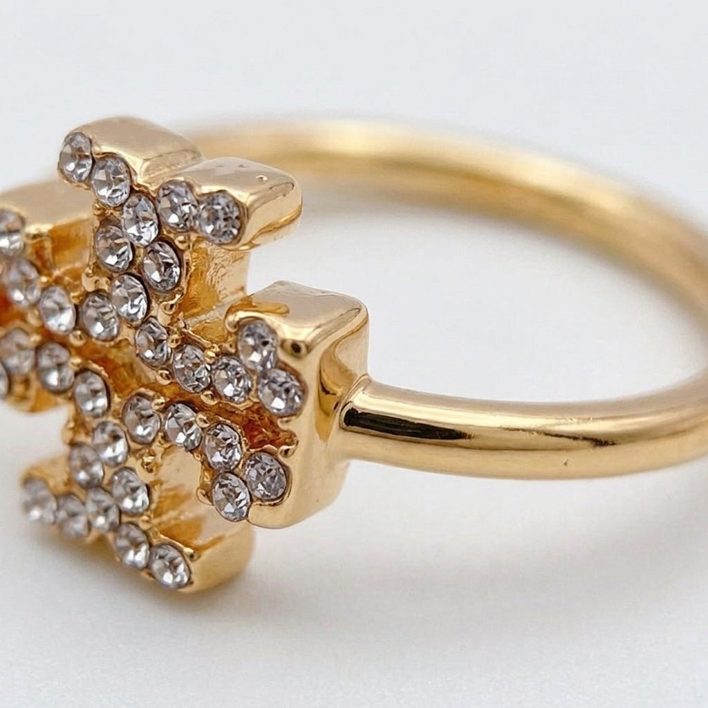 A gold ring featuring a cross-shaped top paved with clear round crystals, shown in a close-up shot against a light gray background.
