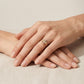A close-up of a person's hands resting on a neutral beige fabric surface, wearing a delicate gold ring with a small cluster of clear stones on the ring finger.