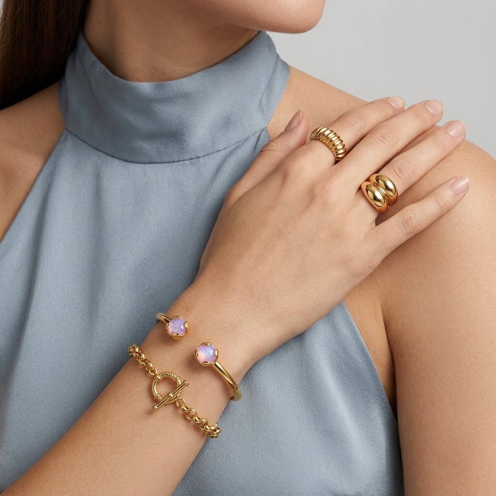 A close-up of a model wearing gold jewelry, including a gold chain bracelet with two round pale pinkish-purple cabochon stones and a gold toggle clasp bracelet, along with gold rings, set against a light blue high-neck top and a neutral gray background.