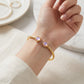 A close-up of a person's wrist wearing a gold-toned open cuff bracelet with two round light purple gemstones, styled with a neutral cream-colored top and a soft marble background.