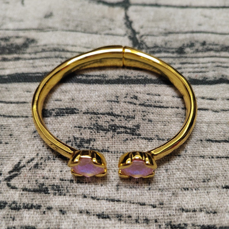 A gold-plated bangle bracelet with two small iridescent pinkish-purple stone accents at the open ends, displayed on a textured gray fabric background.