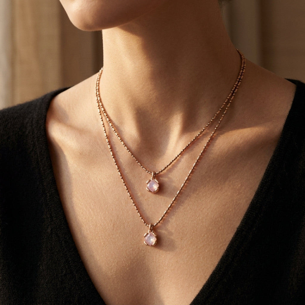 A close-up of a model wearing a double-layered rose gold necklace with small bead chains and teardrop-shaped pale pink gemstone pendants.