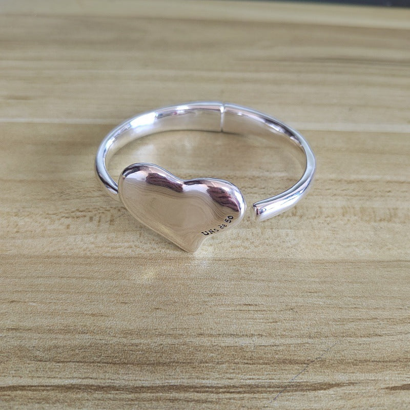 A polished silver open bangle ring with a heart-shaped centerpiece, resting on a light wood grain surface.