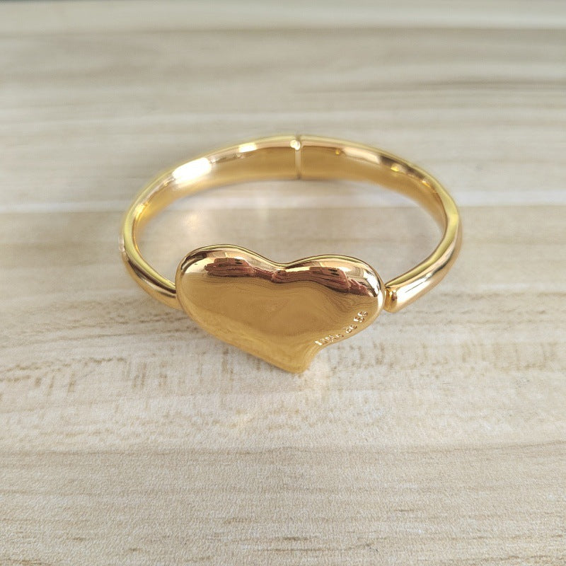 A polished gold-plated adjustable bangle bracelet with a heart-shaped centerpiece, resting on a light-colored wooden surface.