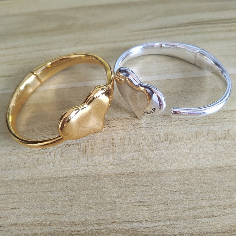 Two heart-shaped bangle bracelets, one in polished gold and one in polished silver, displayed on a light wood grain surface.