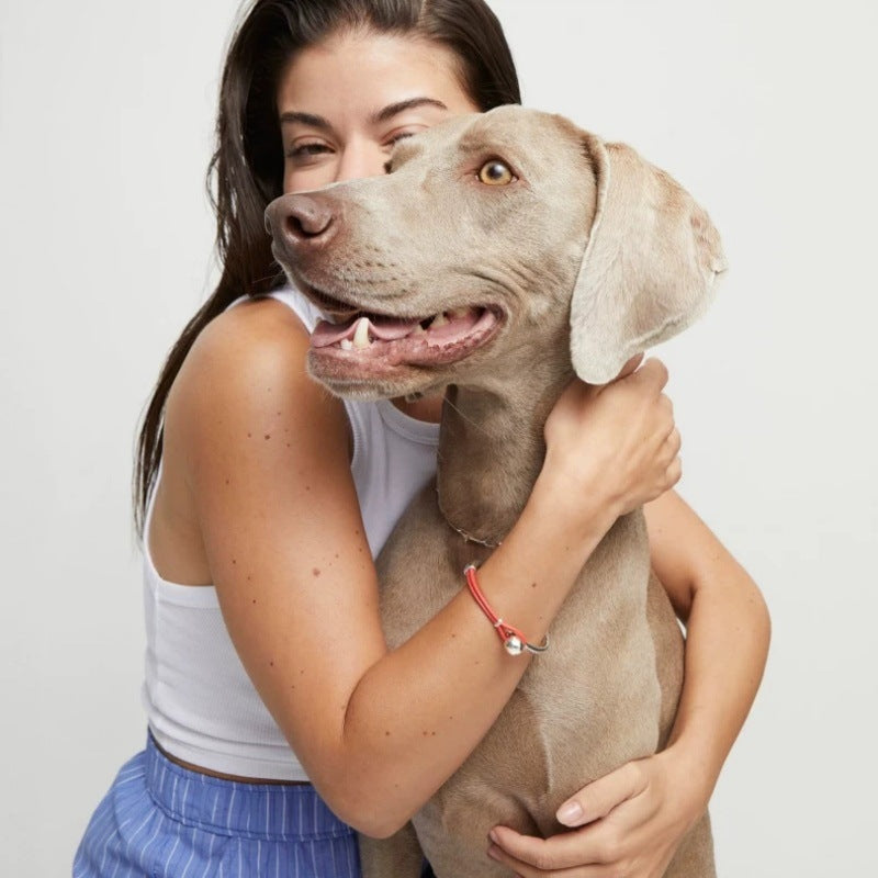 A woman in a white tank top and blue striped pants hugs a large gray dog, wearing a red cord bracelet with a silver charm on her wrist.