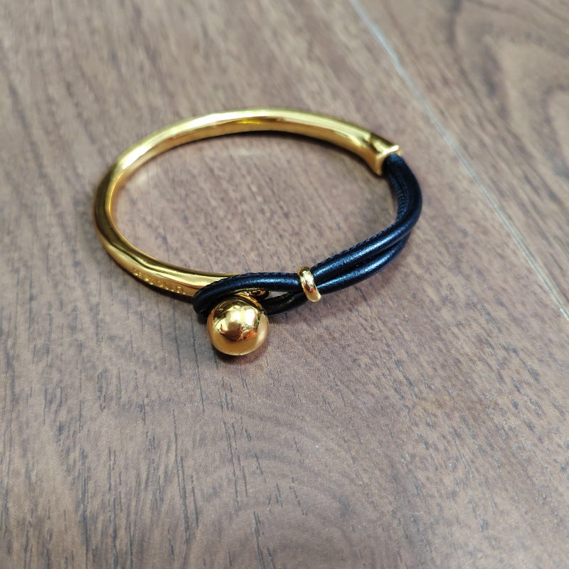 A gold-plated bangle bracelet with a black cord wrap and a small gold bead charm, resting on a brown wooden surface.