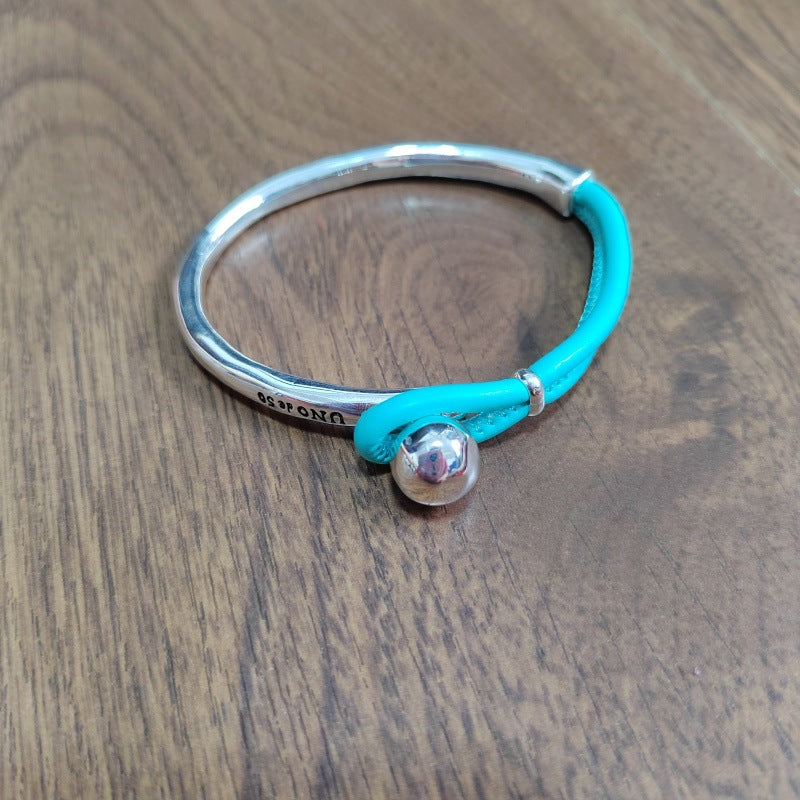 A sterling silver bangle bracelet with a turquoise cord wrap and a small silver bead, resting on a wooden surface.