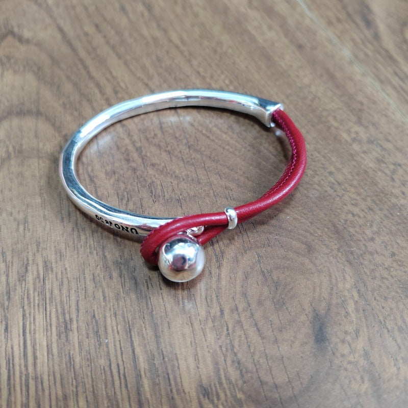 A sterling silver bangle bracelet with a red leather wrap and a small silver bead charm, displayed on a wooden surface.