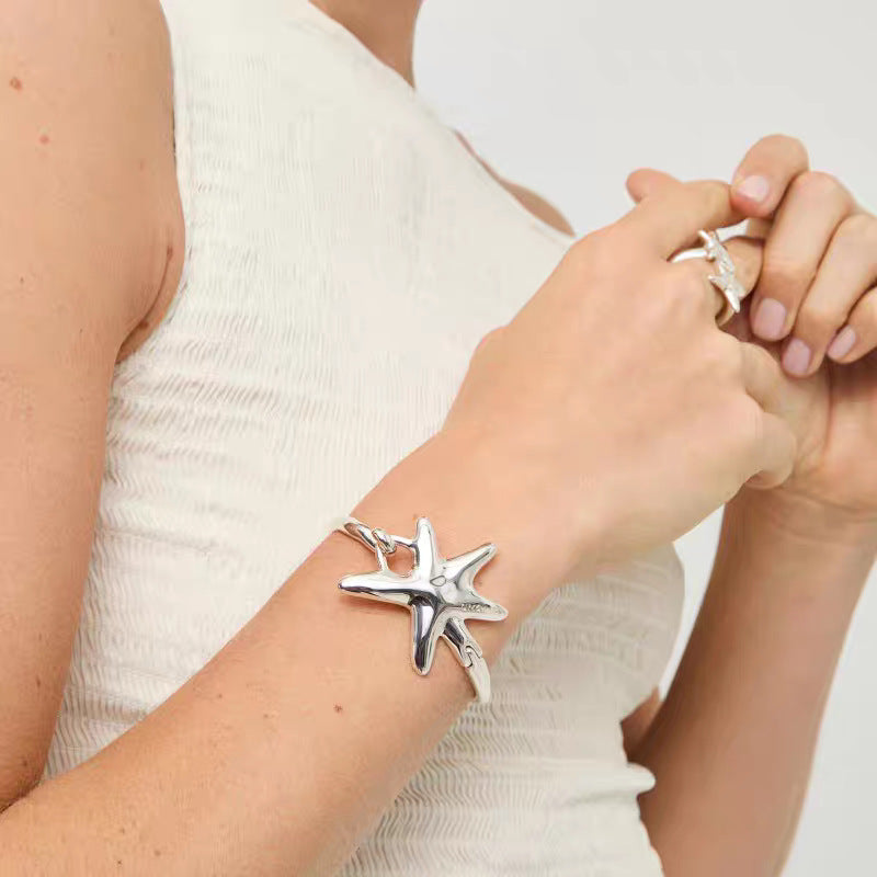 A silver starfish cuff bracelet worn on a woman's wrist, shown in a close-up lifestyle shot with her hands clasped together.