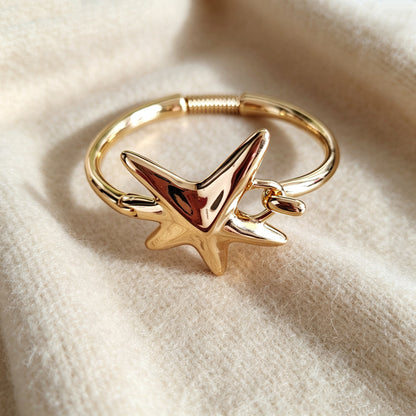 A polished gold-plated starfish ring with a matching starfish charm, resting on a soft, textured cream-colored fabric.