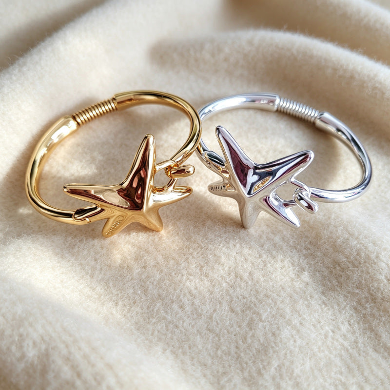 Two open bangle rings with starfish-shaped charms, one in gold and one in silver, displayed on a soft beige fabric background.