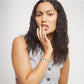 A woman with long, wavy dark hair poses in a light gray ribbed sleeveless top, wearing a silver charm bracelet on her wrist against a plain light gray background.